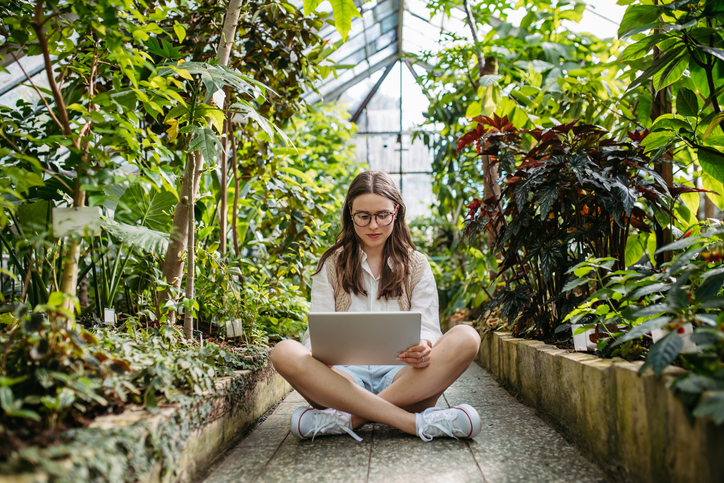 Young woman sitting in a greenhouse and working on the laptop.